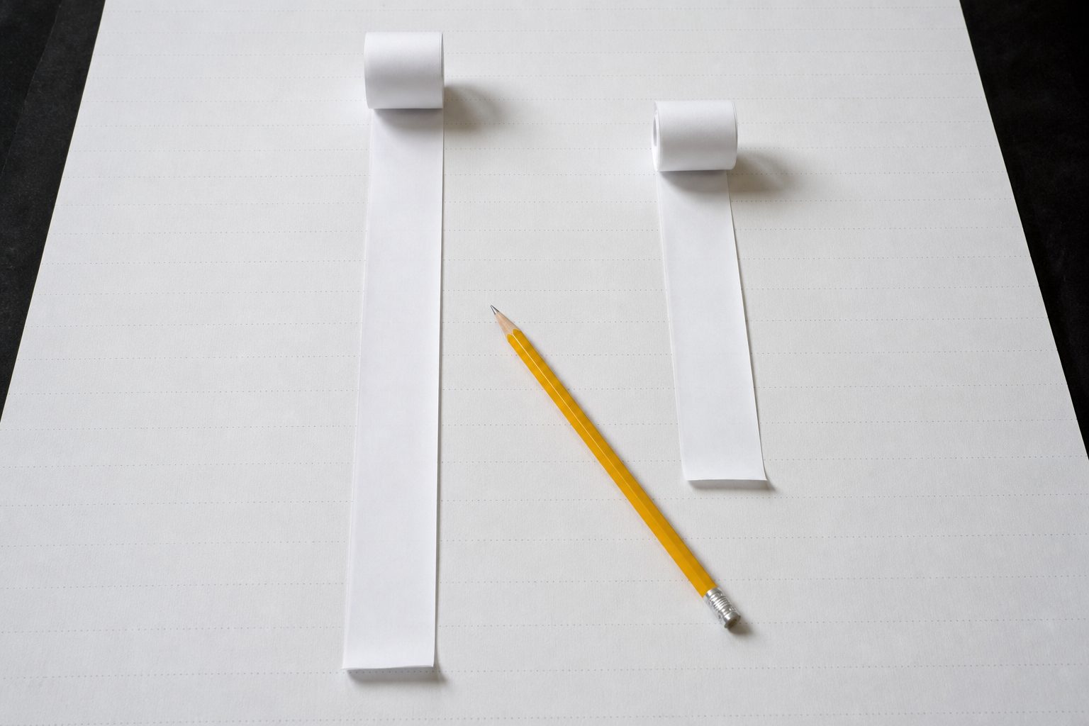 Two adding machine tapes of different lengths laid side by side on white paper with a yellow pencil